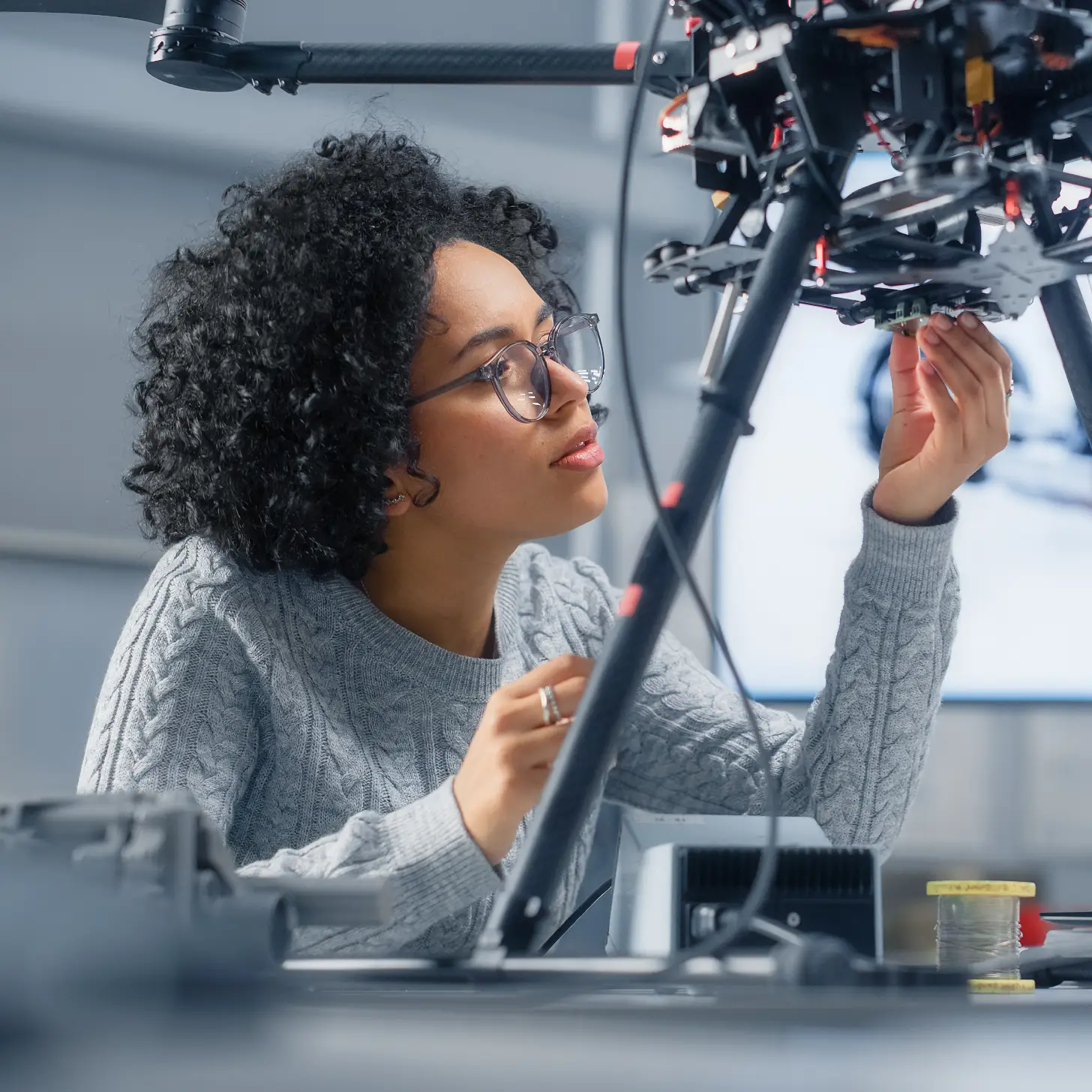 A female engineer in a laboratory examines the underside of a large industrial drone while a technical schematic is visible on a blurred monitor in the background.